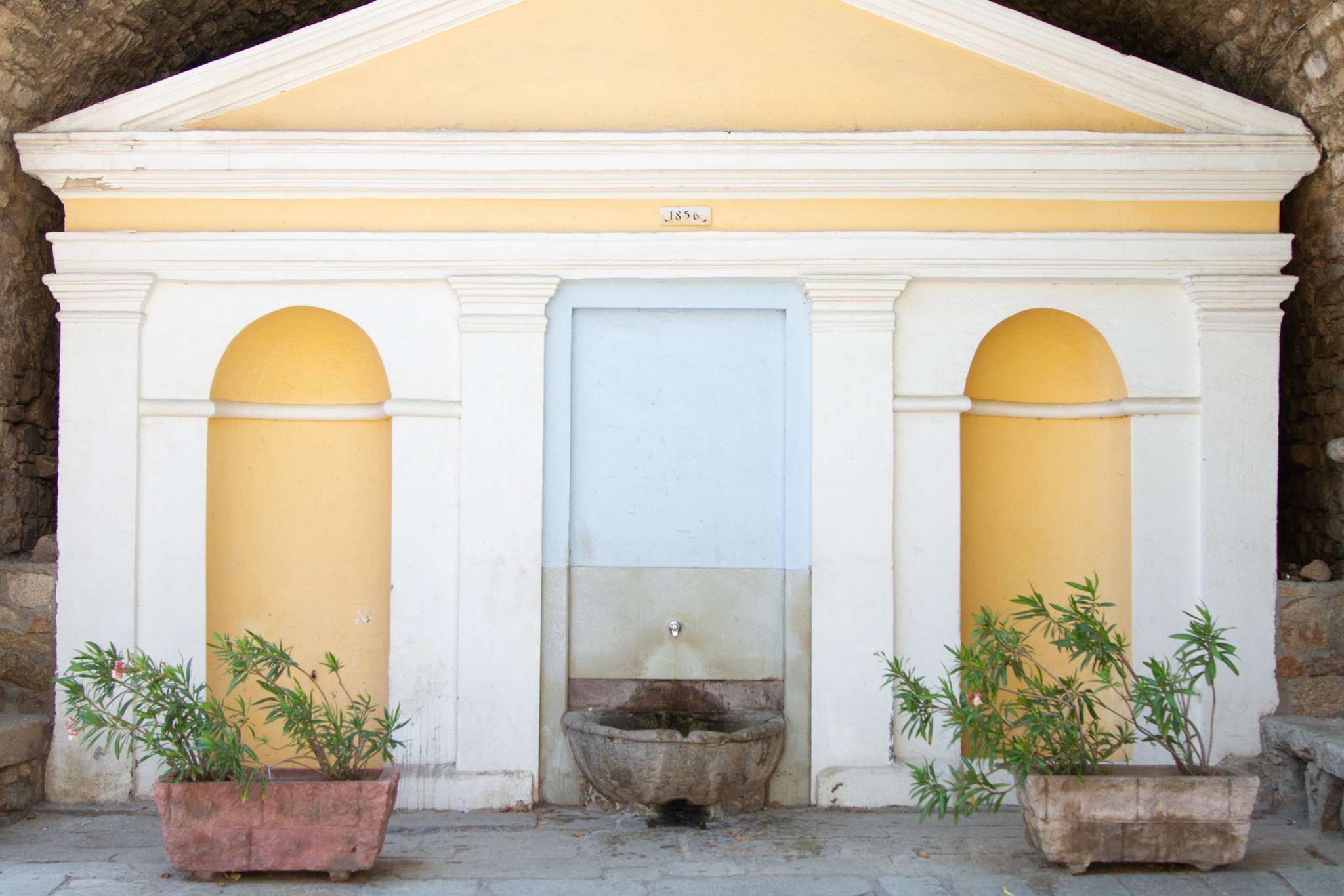 Façade classique blanche avec niches jaunes, colonnes et fontaine centrale entourée de plantes en pot